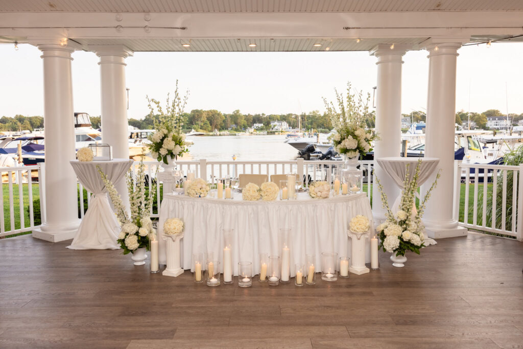 Elegant sweetheart table decorated with white floral arrangements and candlelight on the waterfront pavilion at The Snapper Inn, overlooking boats on the Great South Bay.