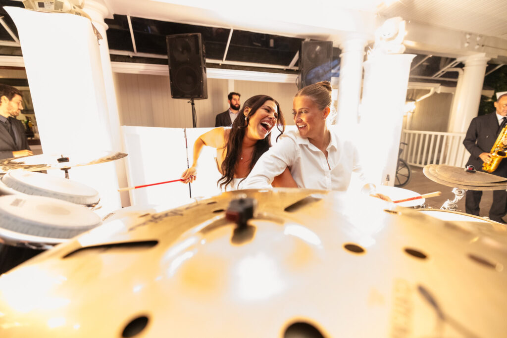 Brides laughing together while playing drums during their lively wedding reception, surrounded by musicians and guests.
