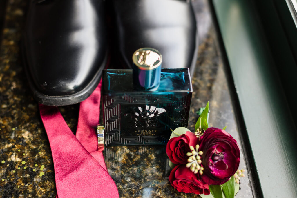 Groom’s wedding day details featuring black dress shoes, red tie, Versace cologne, gold wedding band, and burgundy boutonniere at Westbury Manor.