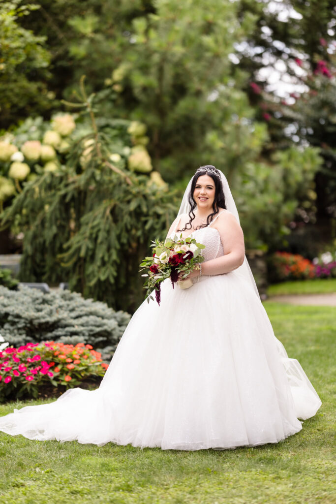 Bride standing in Westbury Manor garden with elegant ballgown and burgundy bouquet, framed by vibrant summer florals and lush greenery.