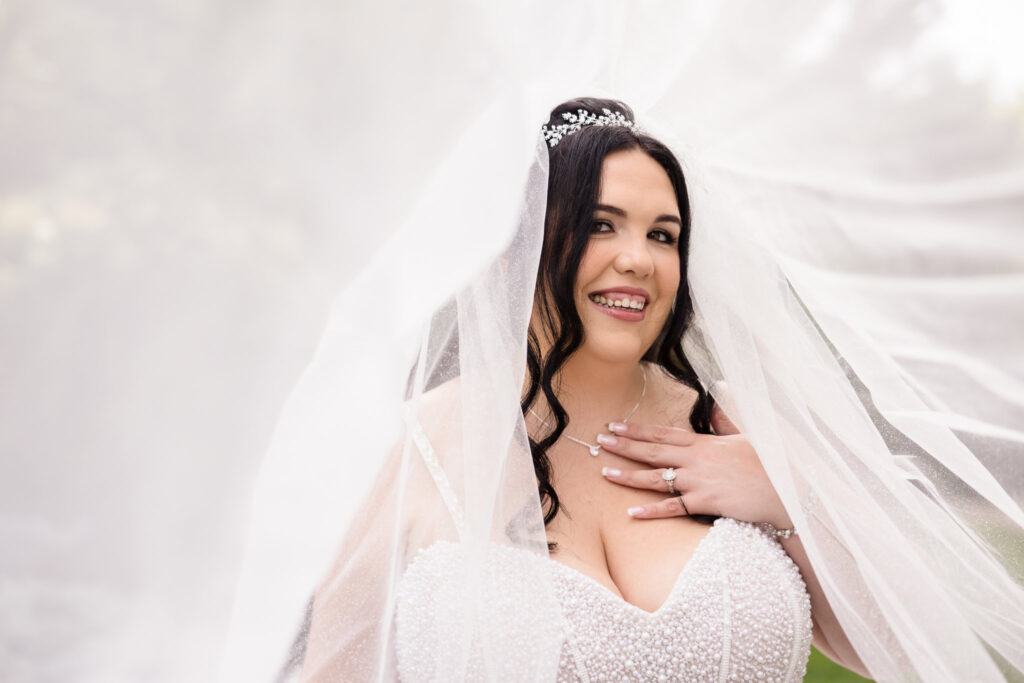 Bride smiling under flowing veil in garden at Westbury Manor, showcasing pearl-beaded gown, diamond engagement ring, and timeless bridal elegance on Long Island.