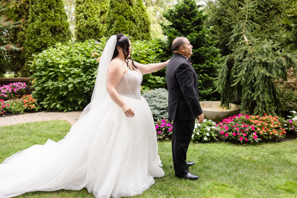 Bride approaching groom for emotional first look in the lush gardens of Westbury Manor, surrounded by vibrant summer florals and evergreen trees.