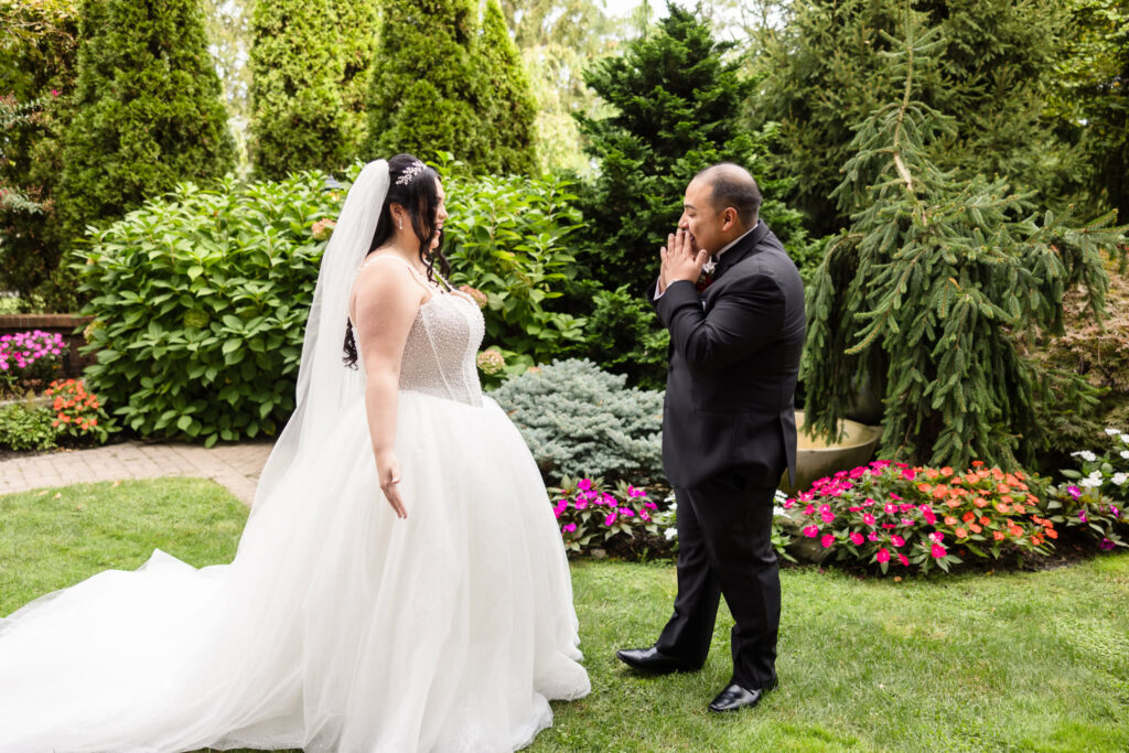 Groom reacts emotionally during first look with bride in Westbury Manor’s garden, surrounded by vibrant flowers and lush greenery.