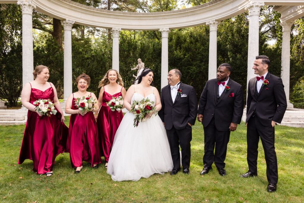 Bridal party laughing together in front of marble columns at Old Westbury Gardens, featuring bride in ballgown and bridesmaids in burgundy satin dresses.
