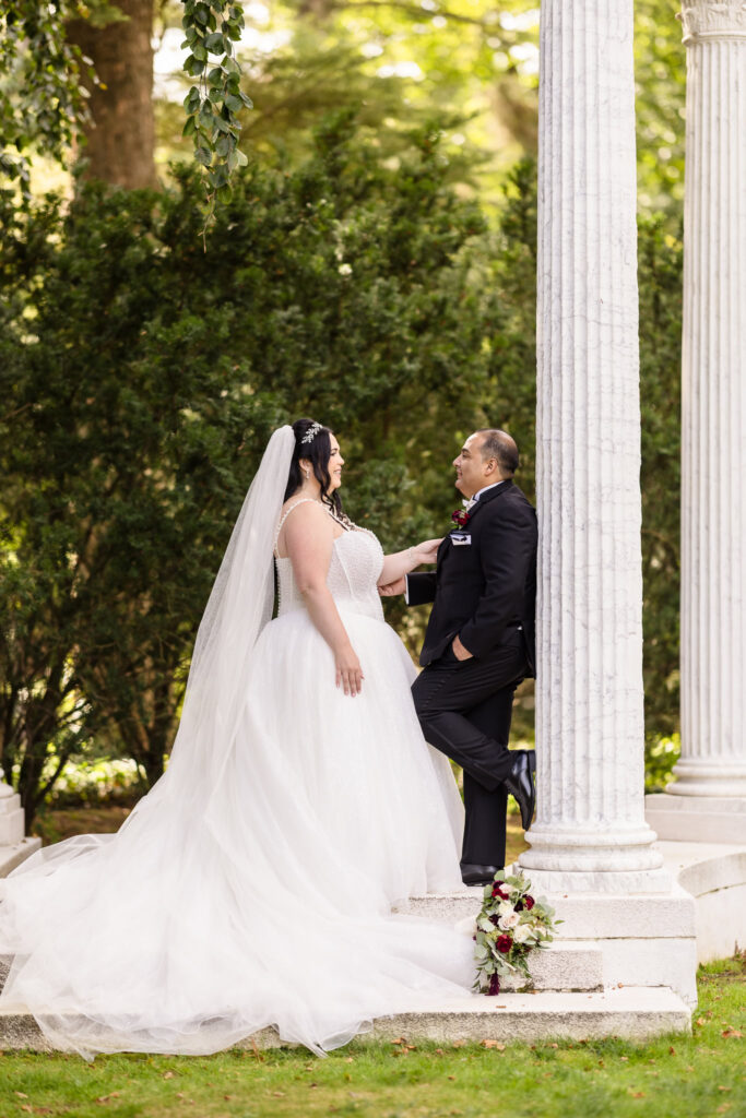 Bride and groom sharing a romantic moment between marble columns at Old Westbury Gardens, with bouquet resting on steps and soft sunlight filtering through trees.