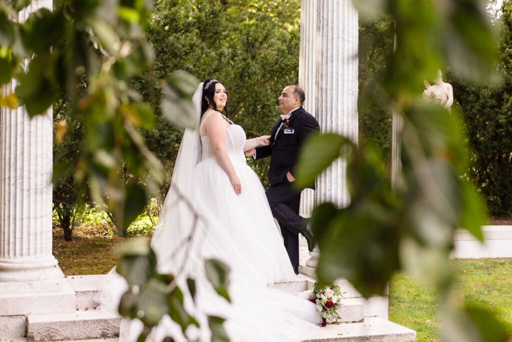 Bride and groom sharing an intimate moment framed by leaves, standing between marble columns at Old Westbury Gardens with bridal bouquet on steps.