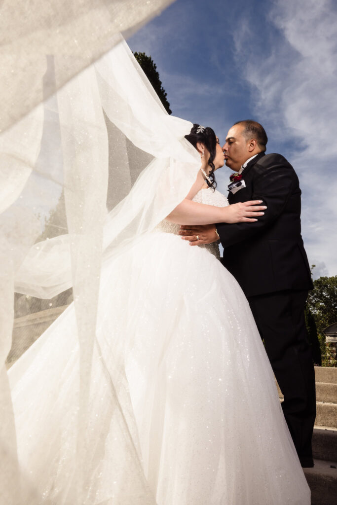 Bride and groom kiss under a flowing veil with dramatic sky backdrop, captured in a cinematic moment of elegance at Old Westbury Gardens.