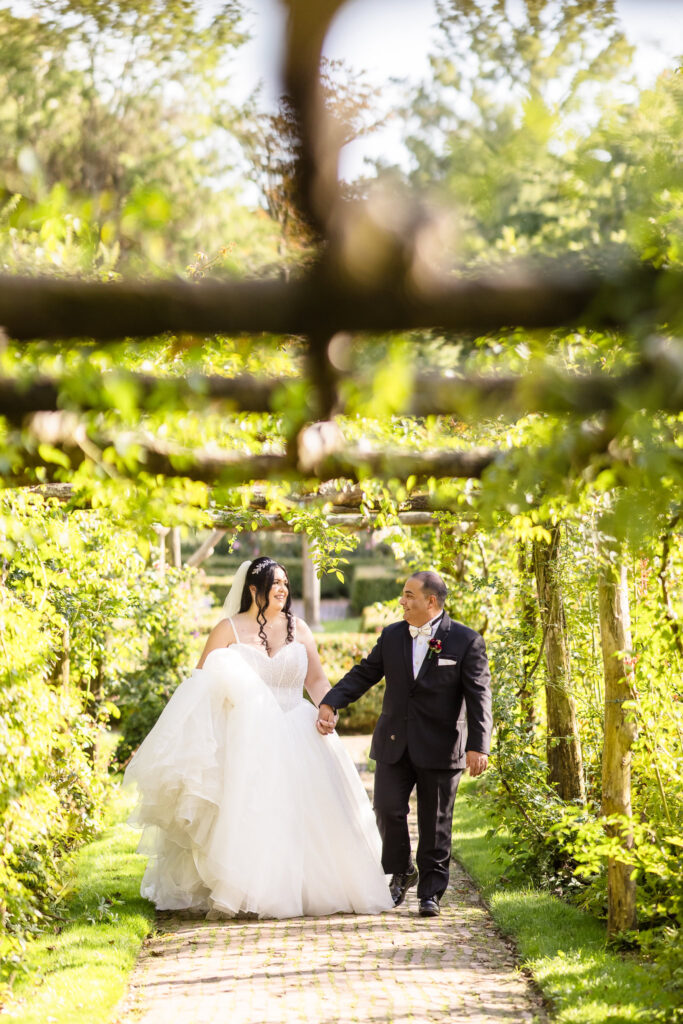 Bride and groom walking hand in hand beneath a sunlit garden trellis at Old Westbury Gardens, radiating joy and timeless elegance on their Long Island wedding day.