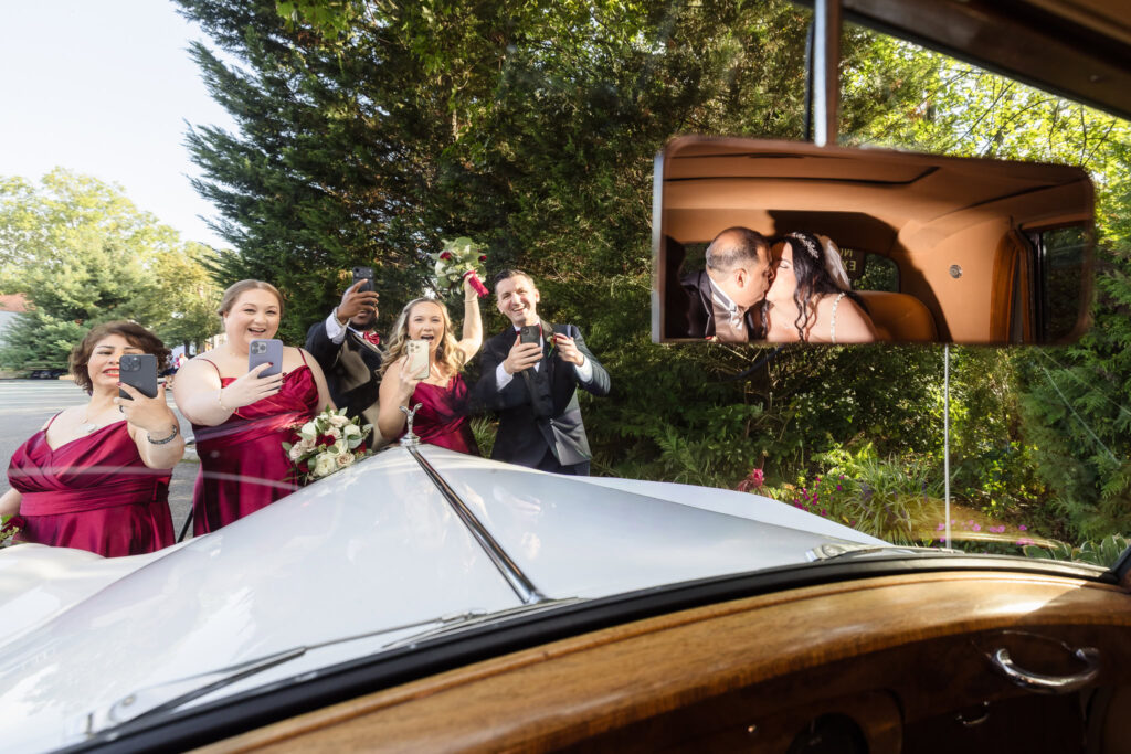 Bridal party cheering outside classic car as bride and groom kiss, reflected in rearview mirror, capturing joyful candid moment after Westbury Manor wedding.