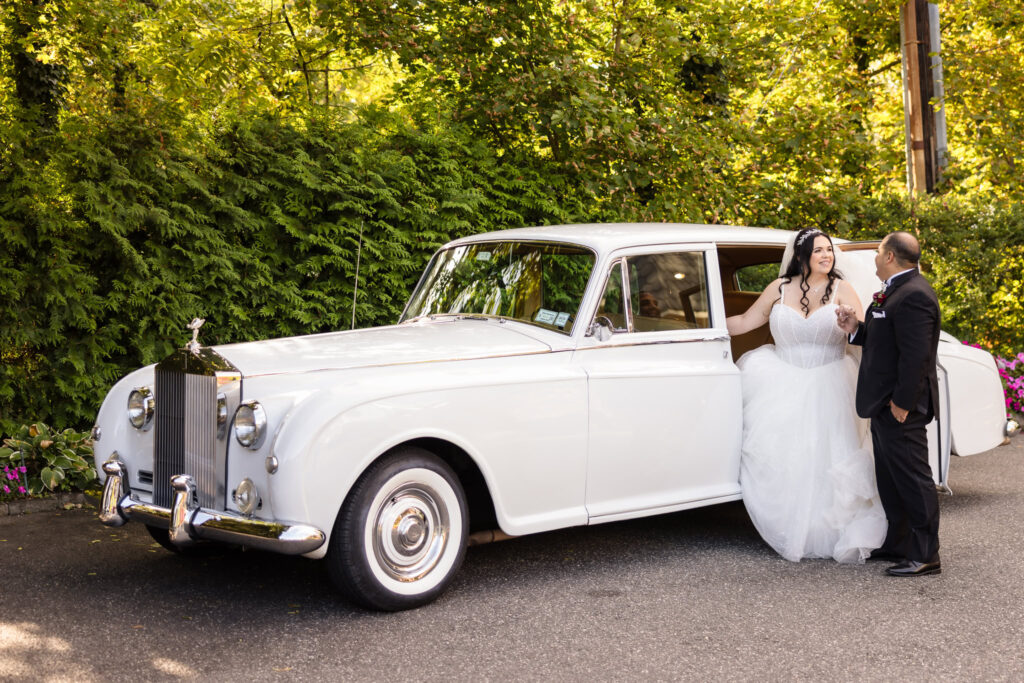 Bride stepping out of classic white Rolls Royce as groom holds her hand, capturing a timeless Westbury Manor wedding moment filled with elegance and charm.
