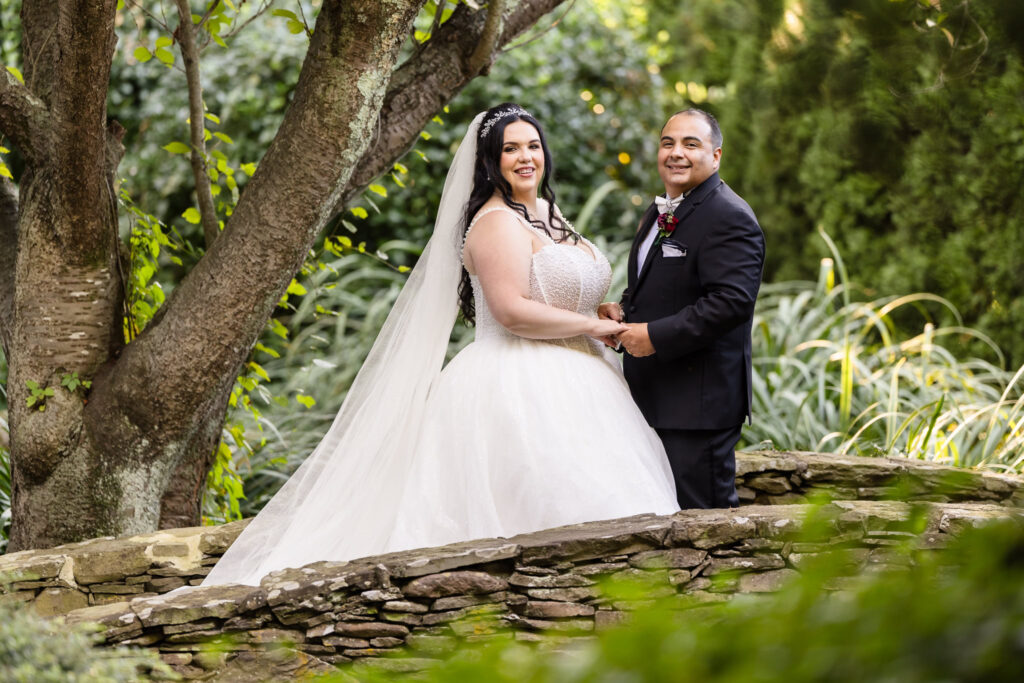 Bride and groom holding hands on a stone bridge surrounded by lush greenery at Westbury Manor, capturing a romantic garden wedding portrait.