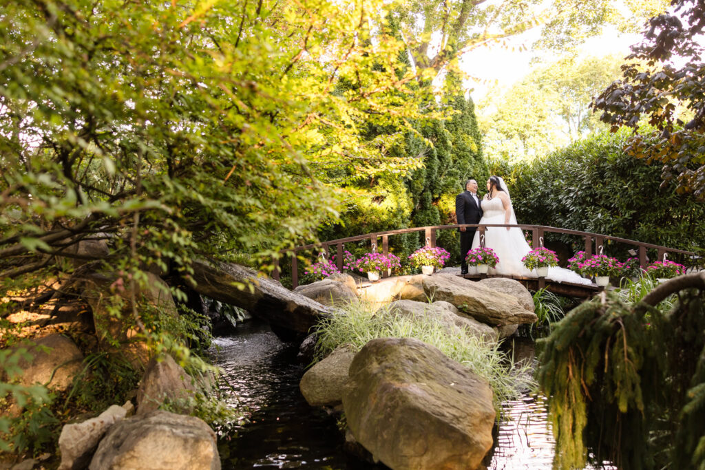 Bride and groom standing on a flower-lined wooden bridge over a tranquil stream at Westbury Manor, surrounded by lush greenery and soft golden light.