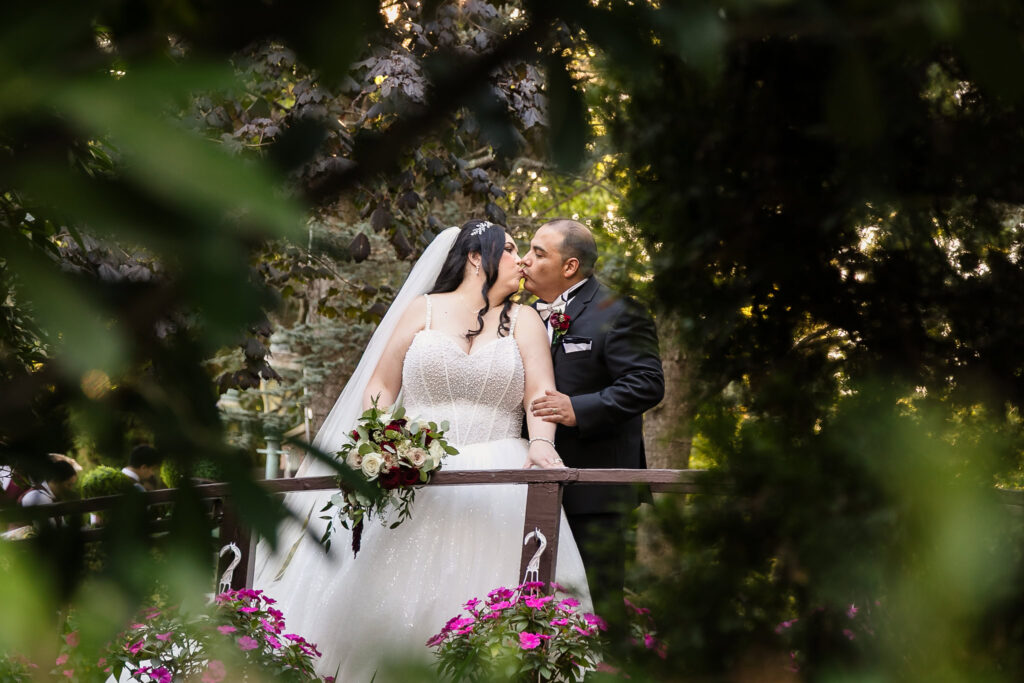 Bride and groom sharing a romantic kiss on a garden bridge framed by flowers and foliage at Westbury Manor.