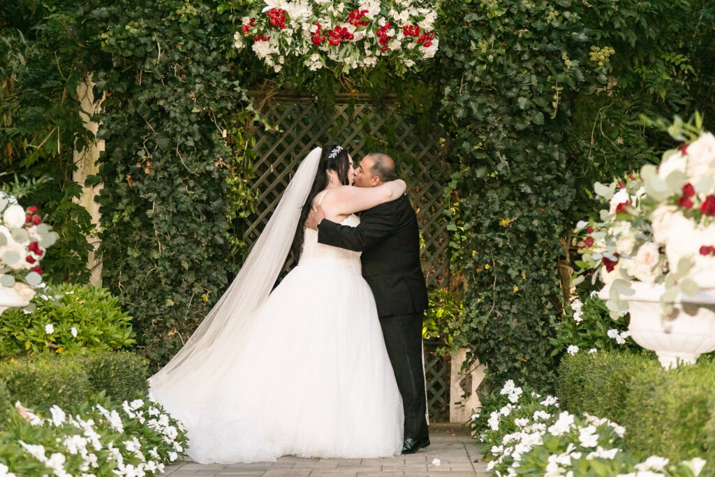 Bride and groom share their first kiss under a lush floral ceremony arch with white and red flowers at Westbury Manor.
