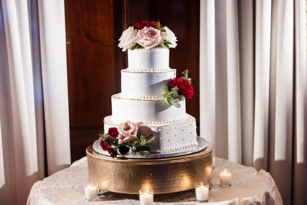 Elegant four-tier white wedding cake adorned with blush and burgundy roses, displayed on a gold stand with votive candles at Westbury Manor.