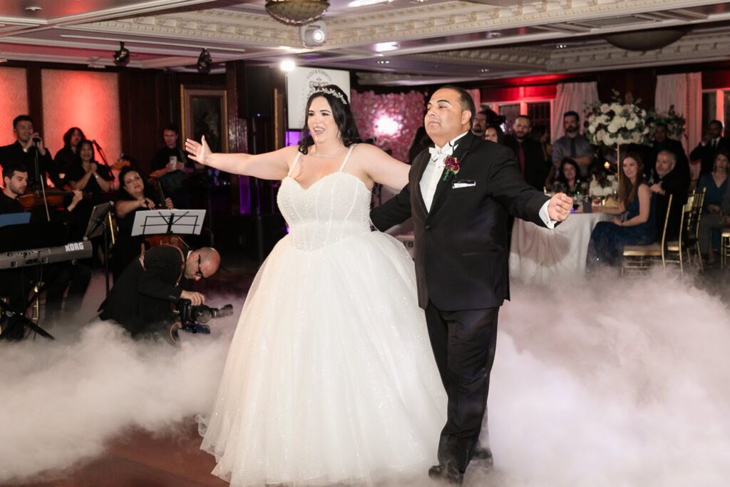 Bride and groom joyfully share their first dance surrounded by fog and live musicians, with guests watching at Westbury Manor’s grand ballroom.