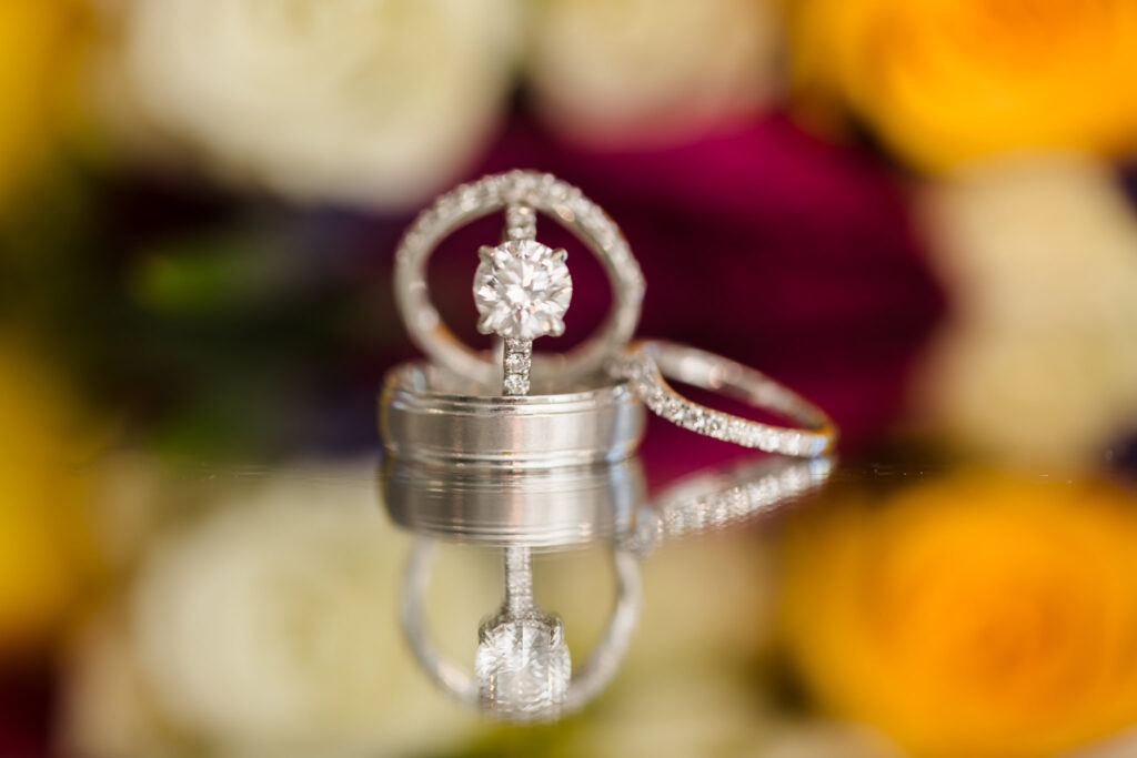 Close-up of wedding rings with diamond engagement ring centered, reflecting on a glass surface with blurred vibrant florals in the background.