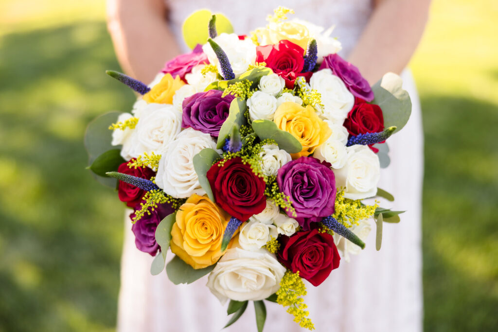 Bridal bouquet with red, purple, yellow, and ivory roses accented by greenery and blue veronica flowers, held against a sunlit vineyard lawn.
