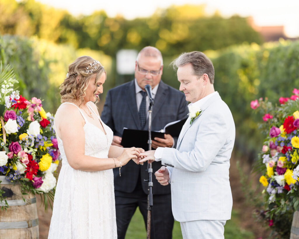 Bride placing the wedding ring on groom’s finger during vineyard ceremony at Baiting Hollow Farm, surrounded by vibrant floral arrangements and lush grapevines.