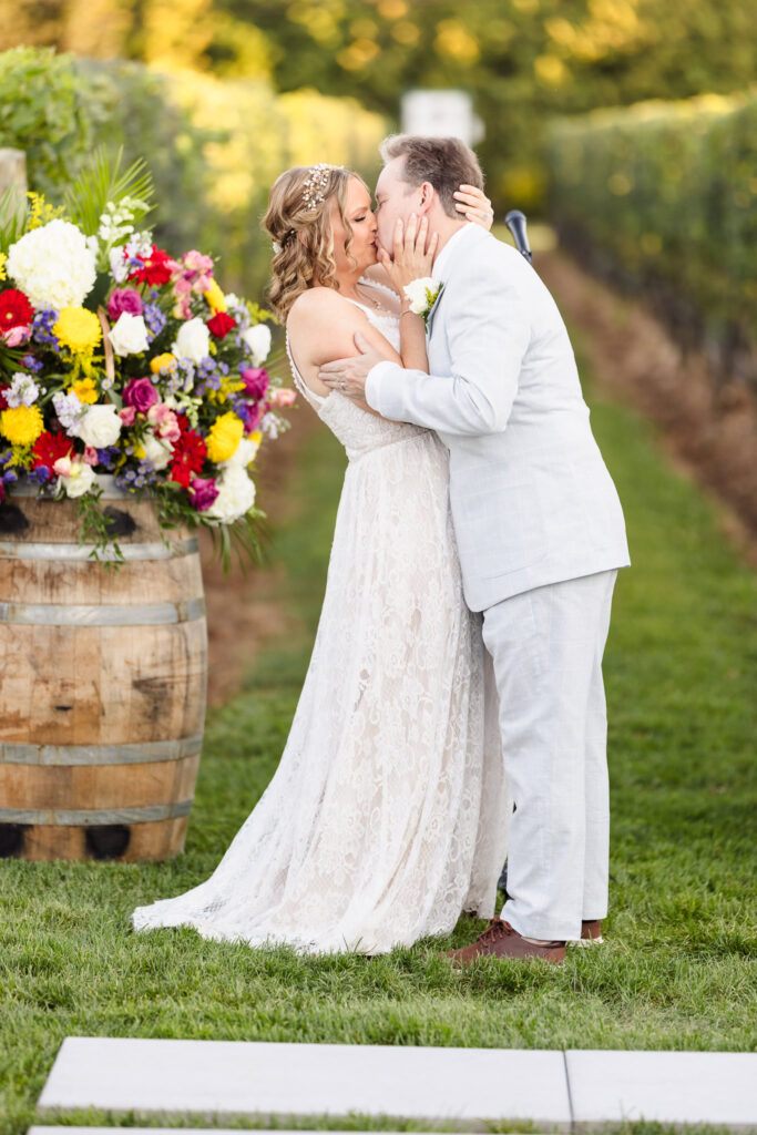 Bride and groom share their first kiss as a married couple during an outdoor vineyard ceremony at Baiting Hollow Farm, framed by colorful floral barrels and summer vines.