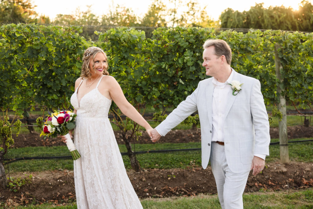 Bride and groom holding hands and smiling at each other while walking through vineyard rows during sunset at Baiting Hollow Farm Vineyard.