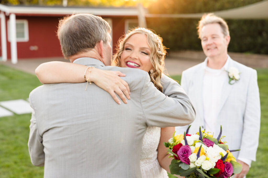Bride embracing her father with a joyful smile during golden hour at Baiting Hollow Farm Vineyard, holding a vibrant bouquet as the groom looks on warmly.
