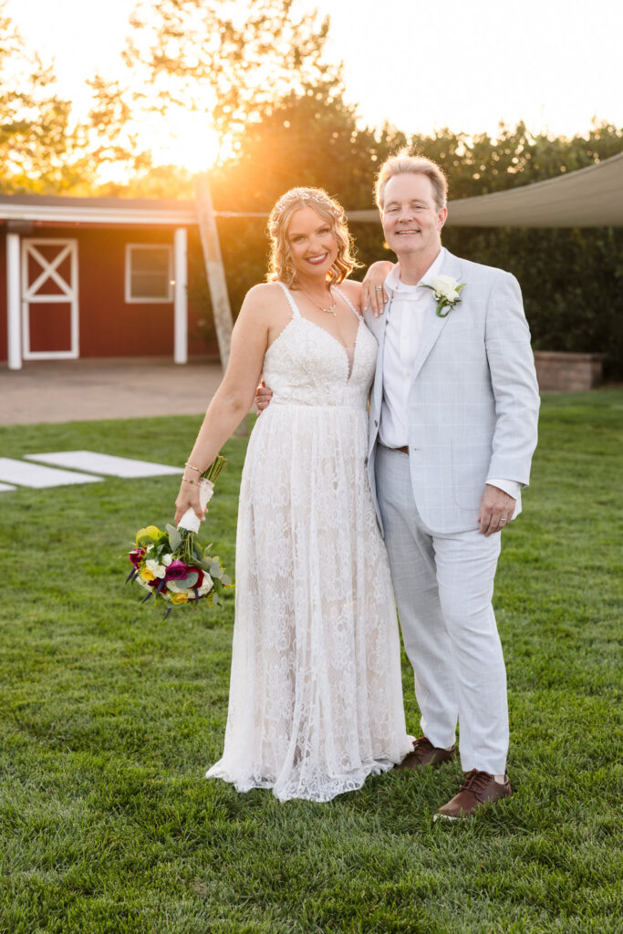 Bride and groom smiling at sunset on the lawn at Baiting Hollow Farm Vineyard, with the bride holding her bouquet and the groom’s arm around her waist.
