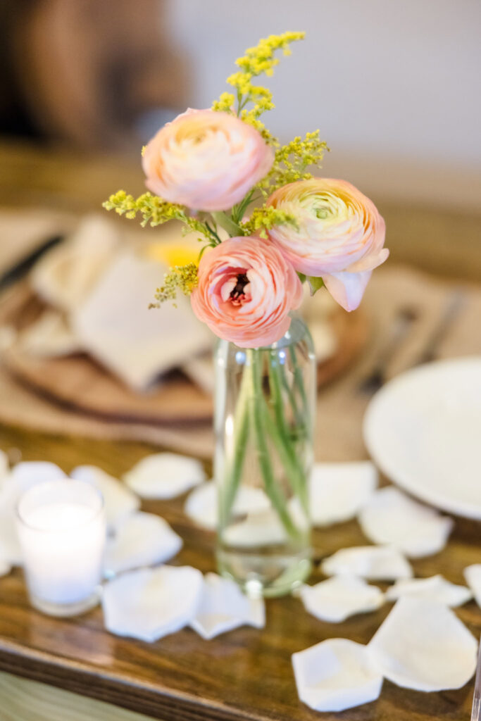 Soft peach ranunculus centerpiece in a clear glass vase with scattered white rose petals and candlelight on a rustic wooden reception table.