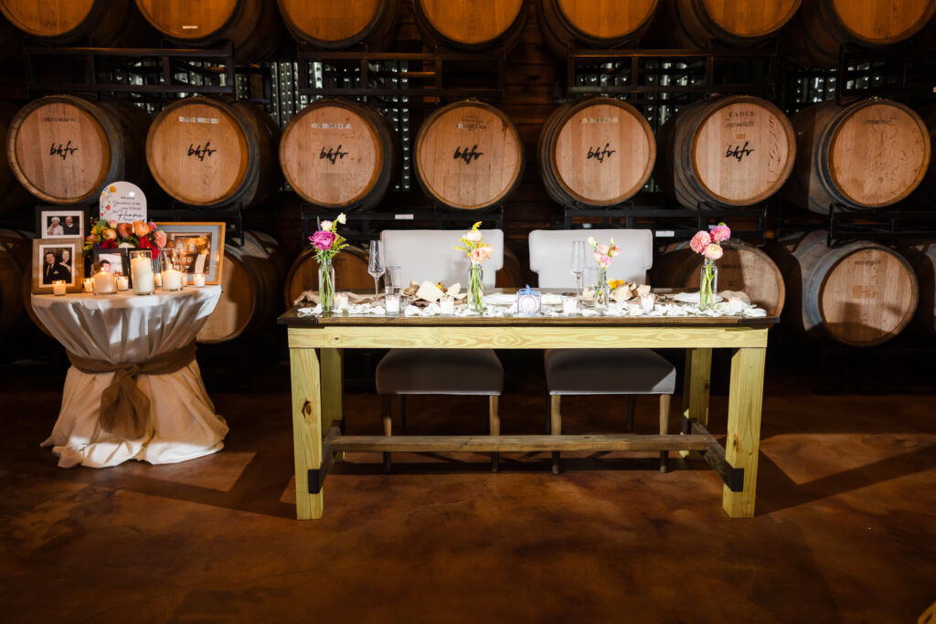 Sweetheart table setup inside The Barrel Room at Baiting Hollow Farm Vineyard, featuring soft floral arrangements, rose petals, candlelight, and a wine barrel backdrop.