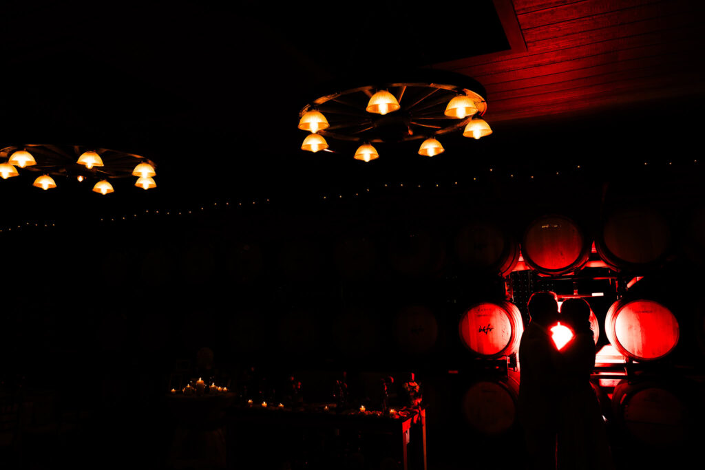 Silhouetted bride and groom sharing a romantic moment in front of wine barrels glowing red in The Barrel Room at Baiting Hollow Farm Vineyard.