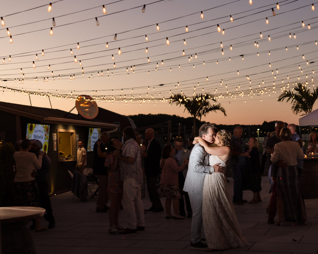 Bride and groom dancing under bistro lights during their outdoor vineyard reception at sunset, surrounded by guests at Baiting Hollow Farm Vineyard.