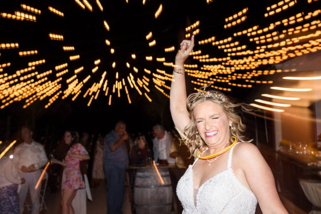 Bride dancing with joy under twinkling string lights during nighttime reception at Baiting Hollow Farm Vineyard, wearing glow necklaces and a radiant smile.