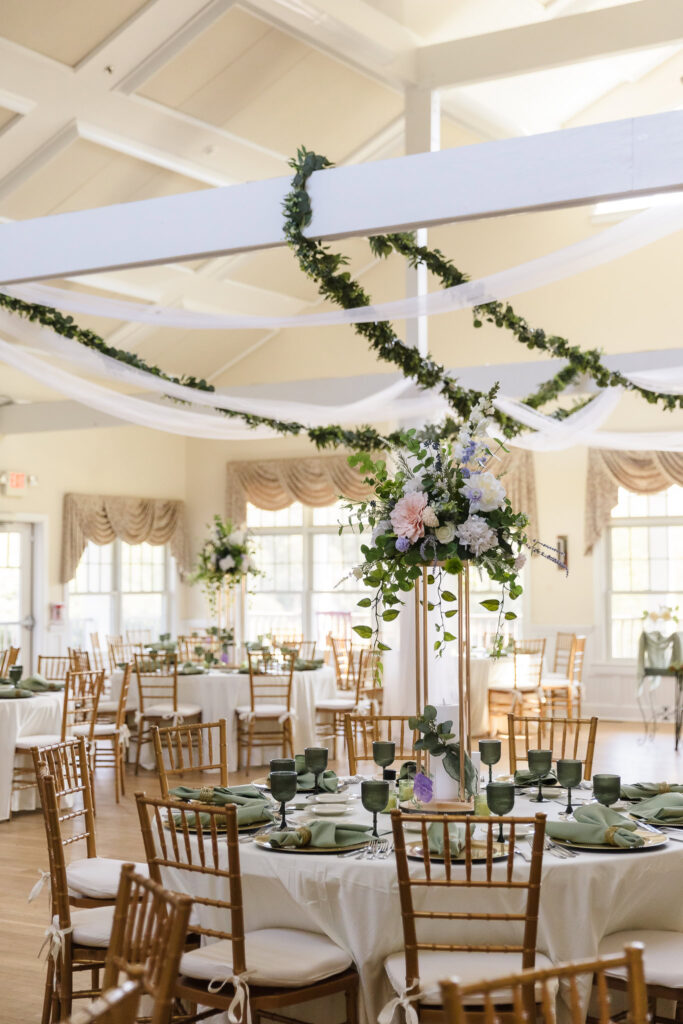 Elegant indoor wedding reception at Camp Pa-Qua-Tuck with tall floral centerpieces, green glassware, and gold chiavari chairs under draped white fabric and greenery garlands.