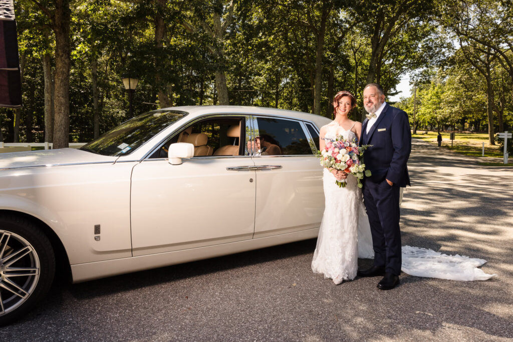 Bride in lace wedding gown holding a pastel bouquet, standing beside a white Rolls-Royce with her father under dappled light at Camp Pa-Qua-Tuck