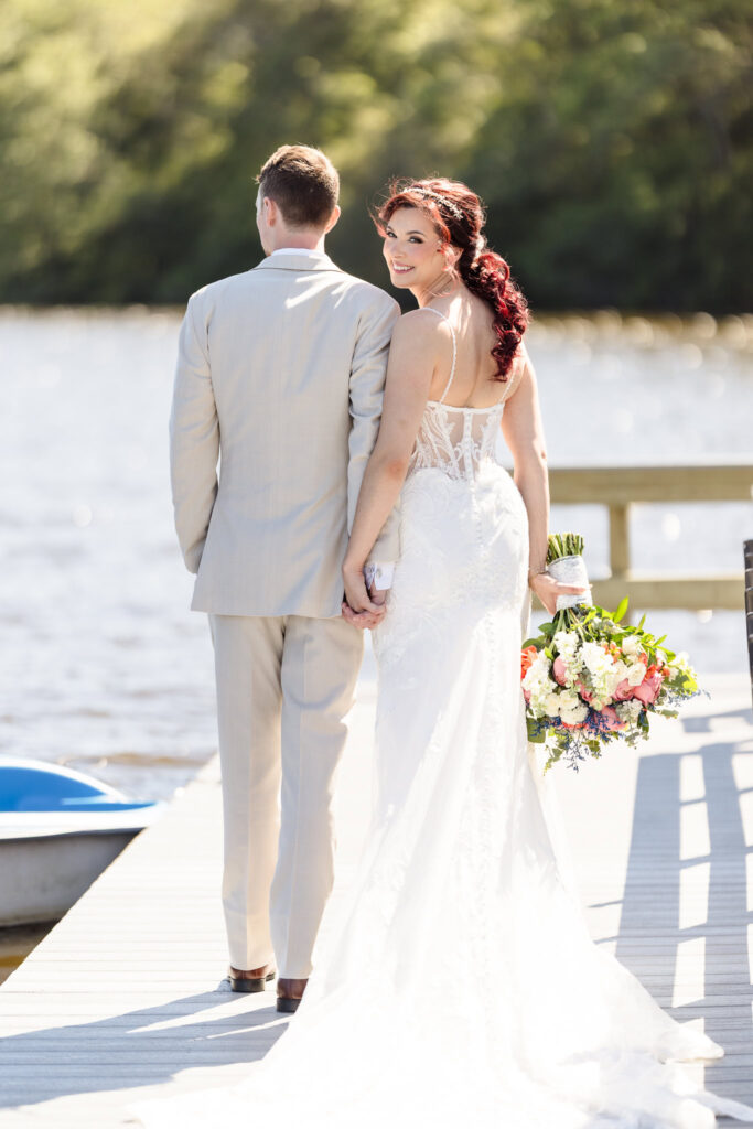 Bride in a lace gown holding a colorful bouquet, smiling over her shoulder while standing hand-in-hand with the groom on the dock at Camp Pa-Qua-Tuck