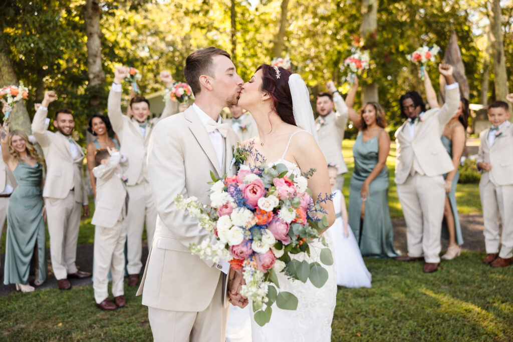 Bride and groom share a kiss while holding a vibrant bouquet, surrounded by a cheering wedding party in sage green and cream attire.
