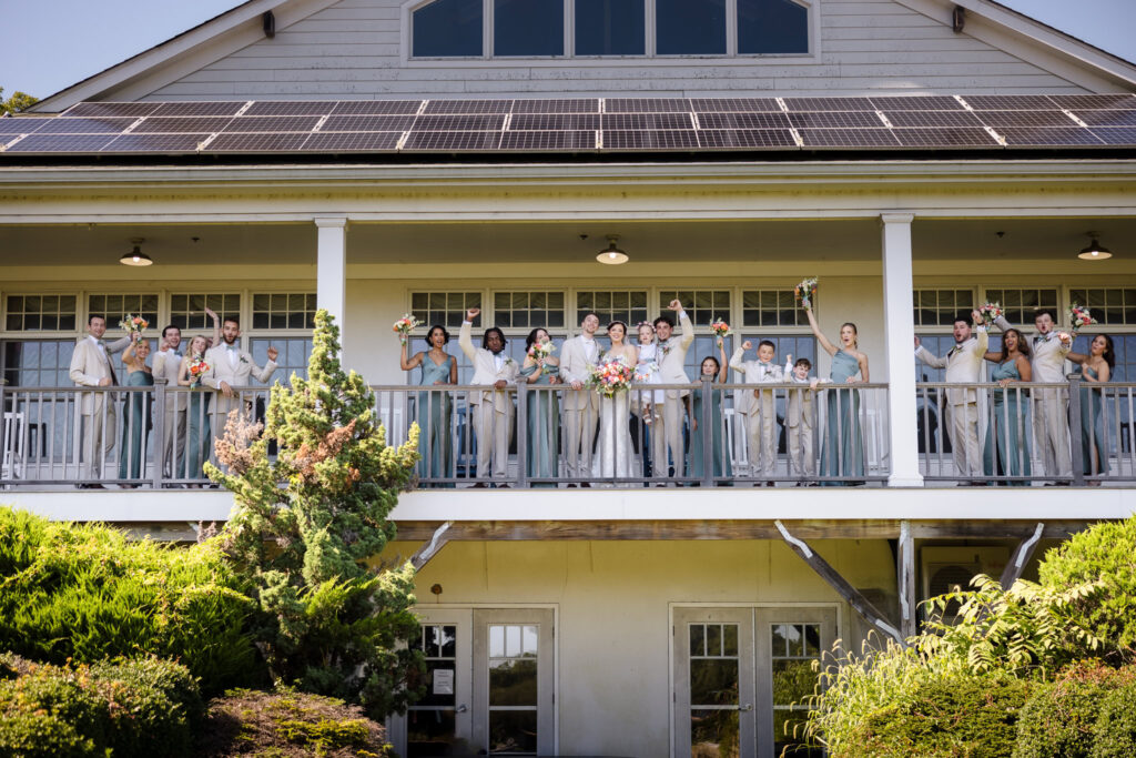 Full wedding party gathered on the upper balcony of Camp Pa-Qua-Tuck’s main building, dressed in sage green and tan, joyfully celebrating with raised bouquets and smiles.