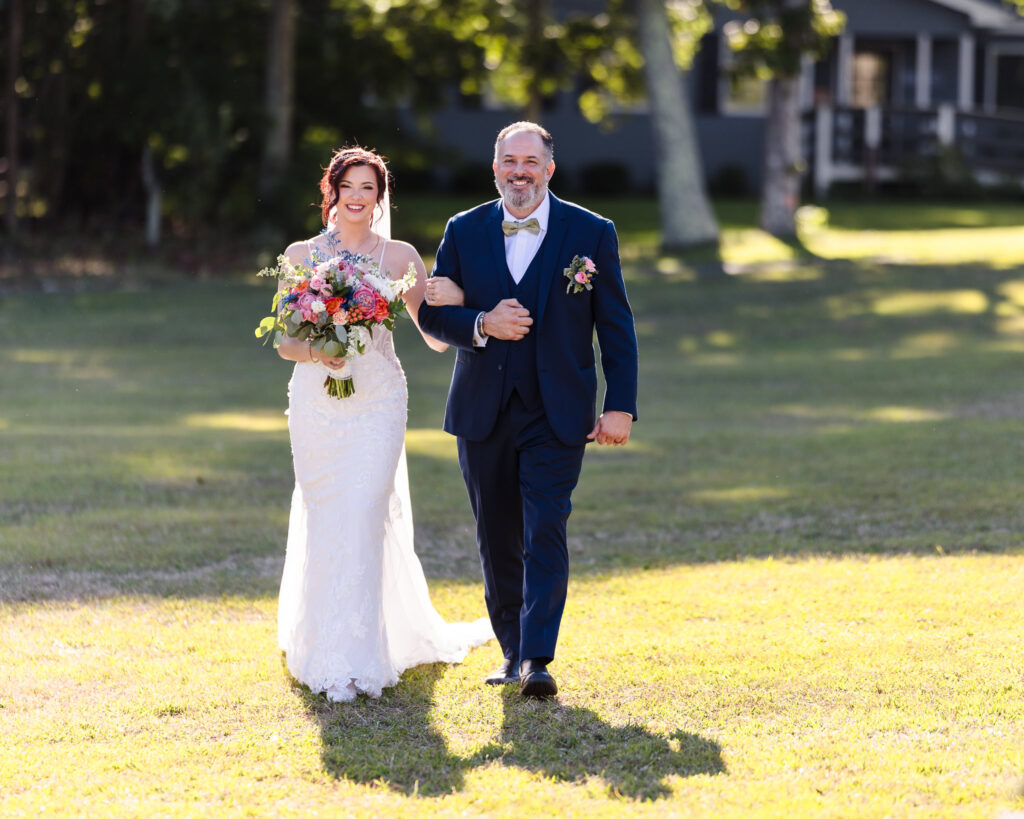 Bride walking arm-in-arm with her father across the lawn, smiling radiantly while holding a colorful bouquet in soft afternoon light.