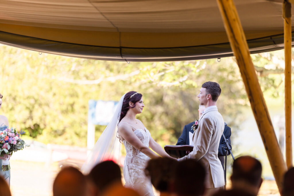 Bride and groom holding hands during their outdoor ceremony under a tent at Camp Pa-Qua-Tuck, exchanging vows with soft natural light filtering through the trees.