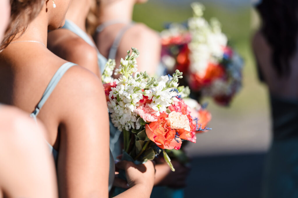 Close-up of bridesmaids in sage green dresses holding vibrant summer bouquets with orange, pink, white, and blue florals.