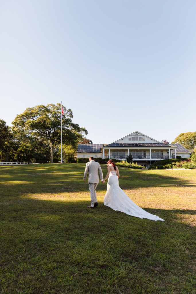 Bride and groom walking hand-in-hand across the open lawn toward the main lodge at Camp Pa-Qua-Tuck, framed by evening light and a clear sky.