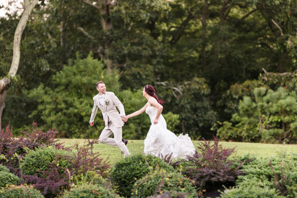 Joyful bride and groom playfully running hand-in-hand through the gardens at Camp Pa-Qua-Tuck, surrounded by vibrant greenery and soft natural light.