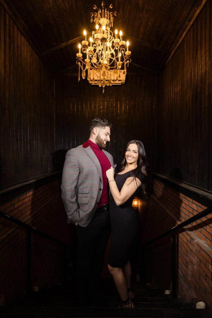 Engaged couple poses beneath a vintage chandelier in the moody brick stairwell of Charlotte’s Speakeasy during their Long Island engagement session.