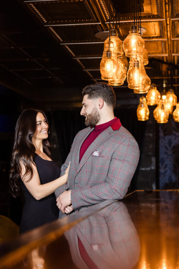 Couple shares a warm gaze under golden pendant lights at Charlotte’s Speakeasy bar during their intimate Long Island engagement session.