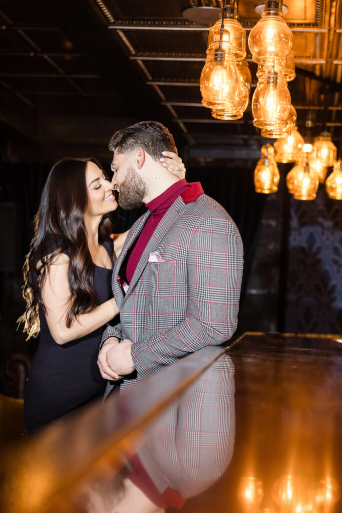 Romantic moment as couple leans in for a kiss beside the bar at Charlotte’s Speakeasy, bathed in vintage light during their Long Island engagement session.