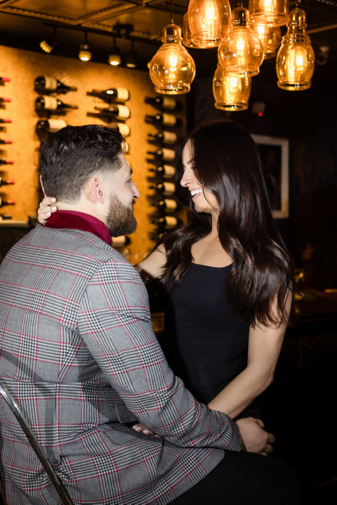 Engaged couple shares a playful smile seated at the bar inside Charlotte’s Speakeasy, surrounded by warm lighting and a wall of vintage wine bottles.