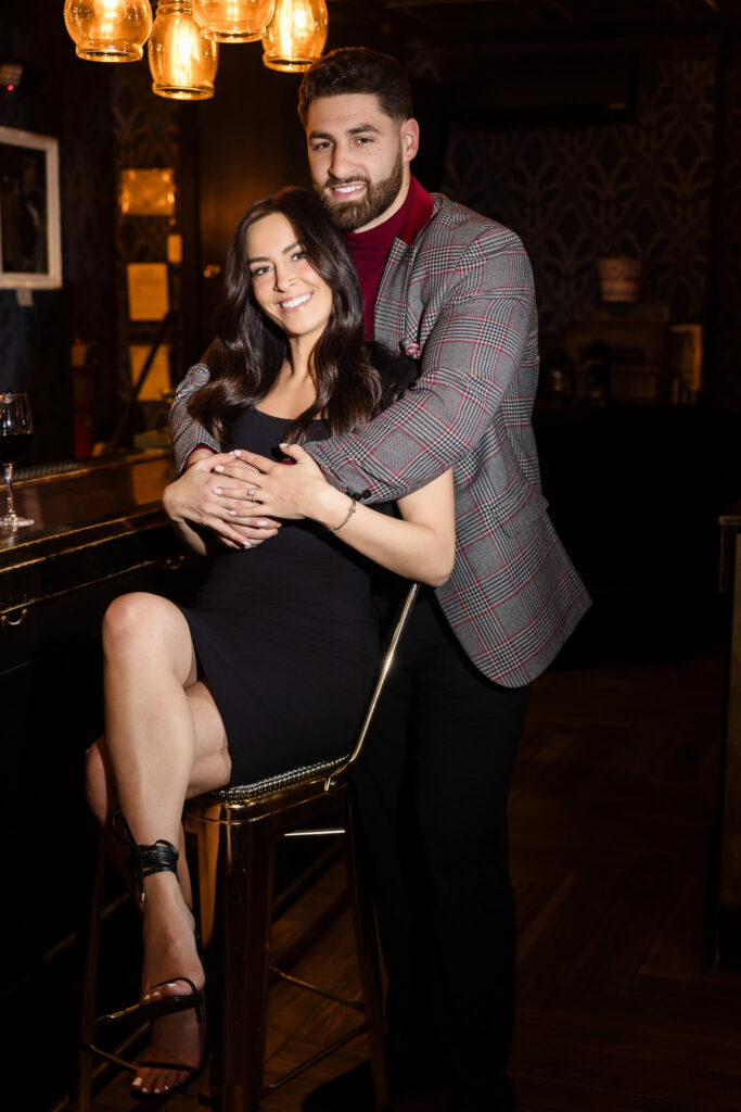 Groom-to-be embraces his fiancée from behind as she smiles at the bar during their Charlotte’s Speakeasy engagement session on Long Island.