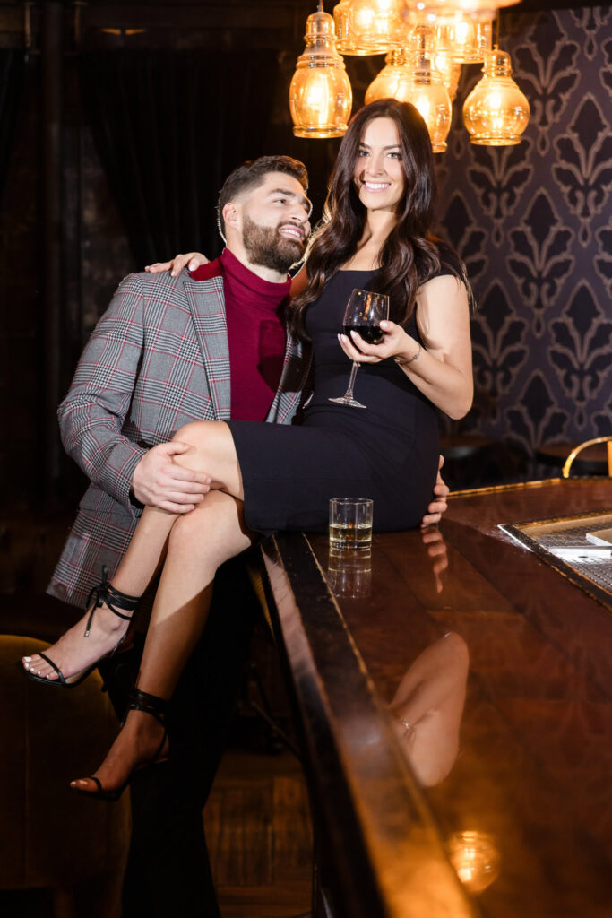 Engaged couple shares a playful moment at the bar in Charlotte’s Speakeasy, with the bride-to-be smiling and holding a glass of wine during their moody Long Island session.