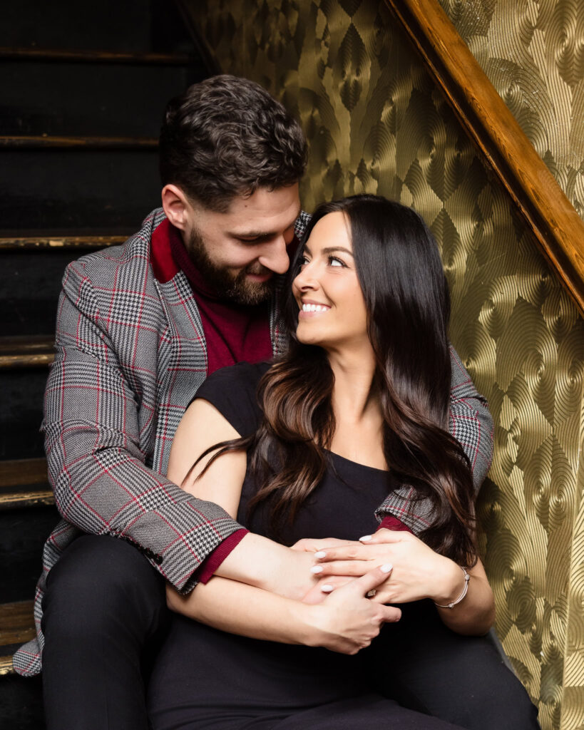 Couple sits close on the vintage staircase at Charlotte’s Speakeasy, sharing a cozy embrace against gold-patterned wallpaper during their Long Island engagement session.