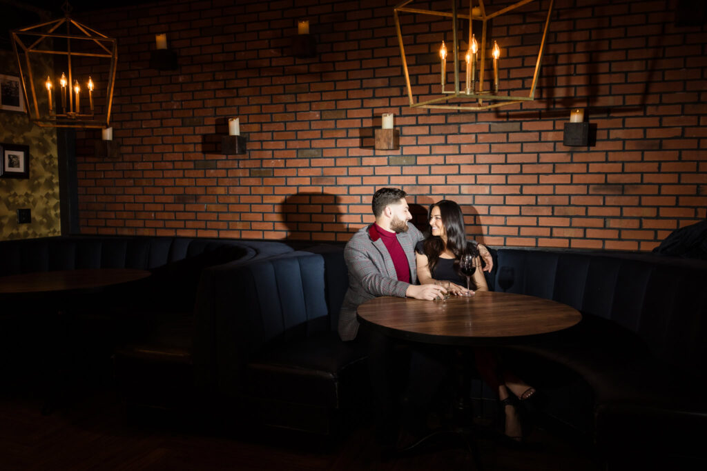 Couple shares a quiet moment over drinks in a cozy booth at Charlotte’s Speakeasy, framed by brick walls and candlelight during their Long Island engagement session.